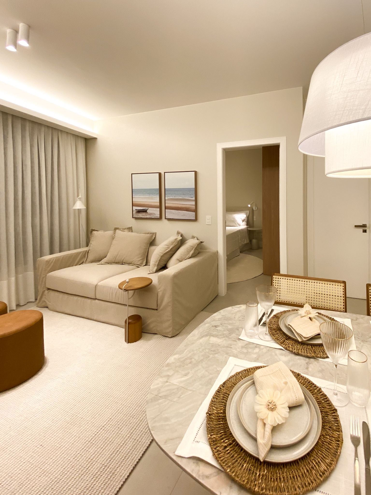 Sala de estar aconchegante em tom bege com sofá, obras de arte, mesa de jantar para dois e uma porta para outro cômodo.