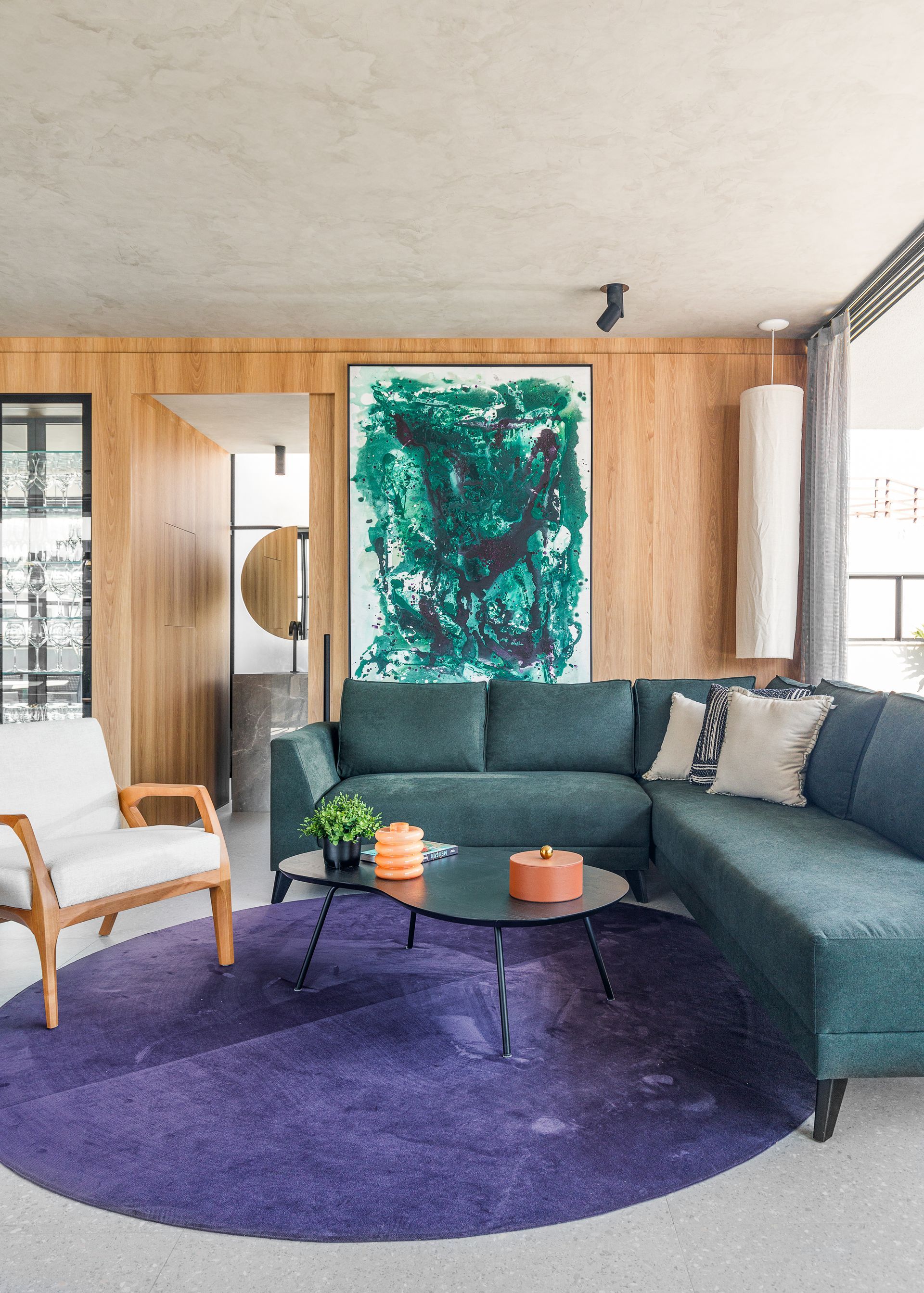 Sala de estar com sofá azul esverdeado, tapete roxo, painéis de madeira, grande pintura abstrata e poltrona branca.
