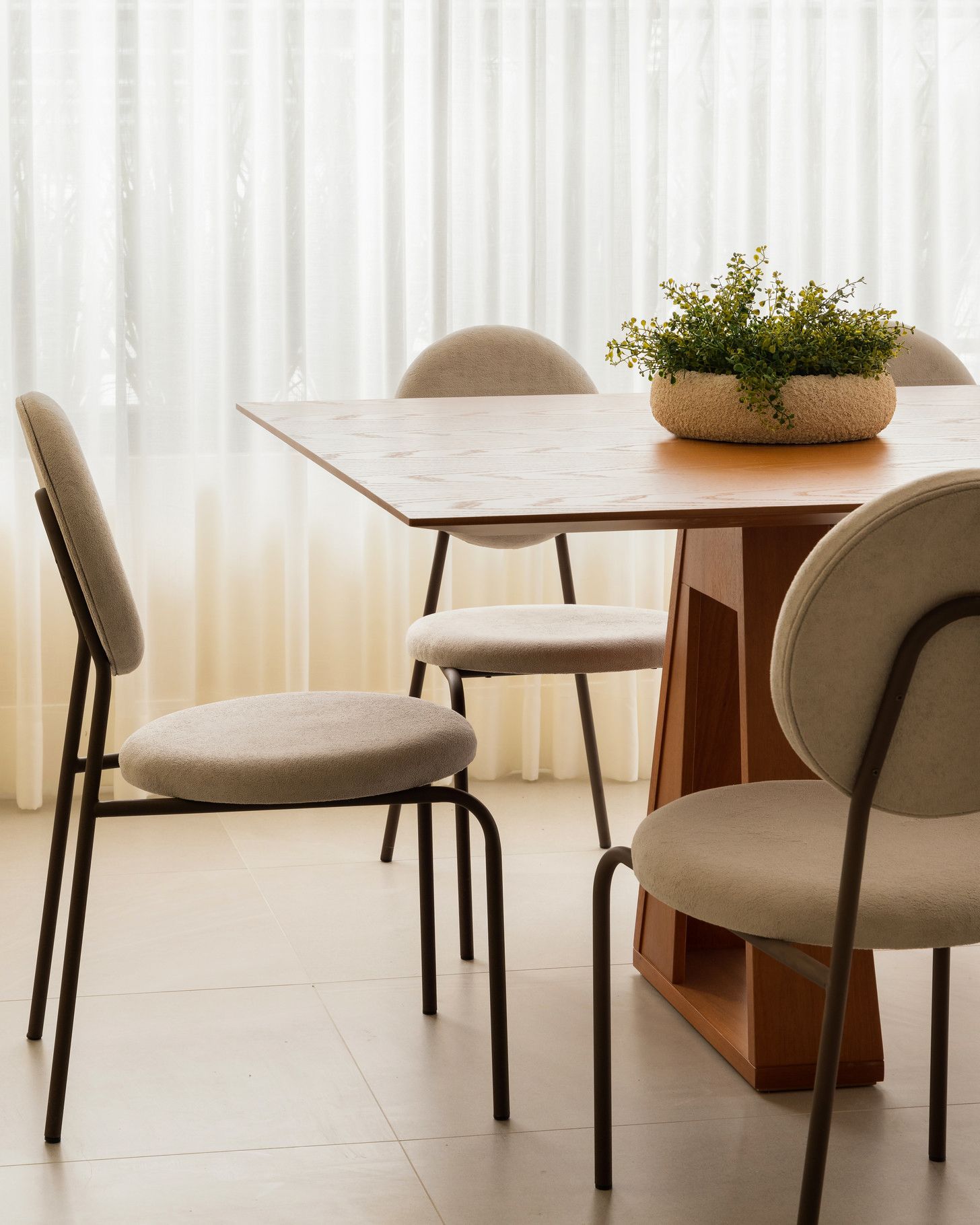 Mesa de jantar com quatro cadeiras bege, uma planta em vaso e cortinas transparentes.