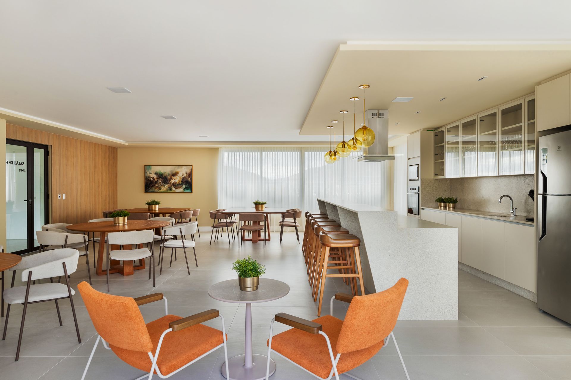 Área comum de conceito aberto com mesas, cadeiras, cozinha e duas poltronas laranja em primeiro plano.