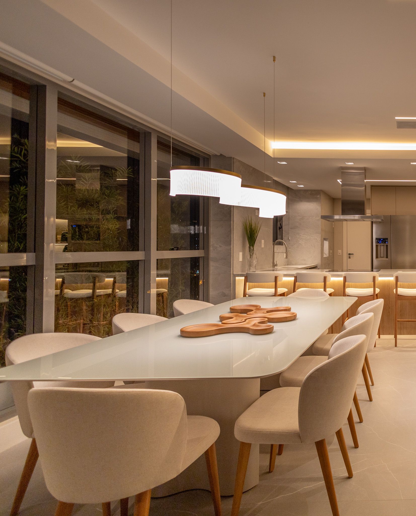 Sala de jantar com mesa comprida, cadeiras creme e luminárias pendentes. Cozinha e janelas ao fundo.