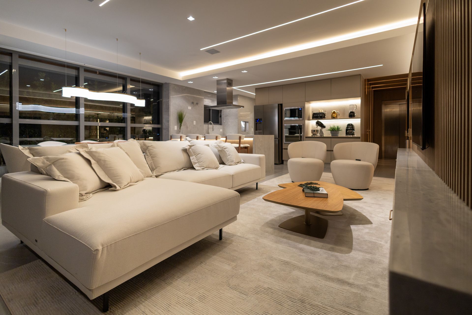 Sala de estar moderna com sofá bege, tapete e mesa de centro. Cozinha ao fundo.