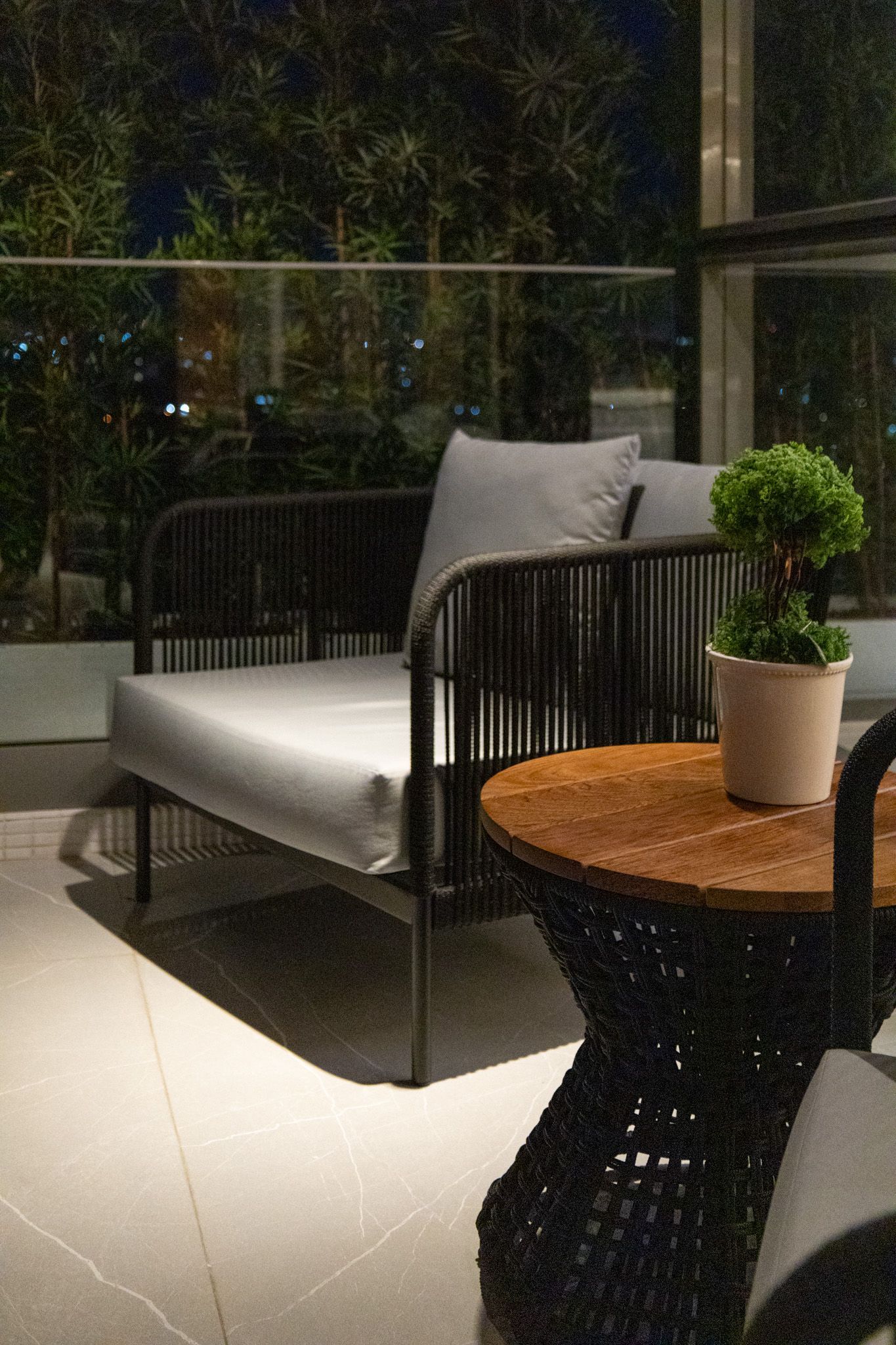 Área de estar na varanda com sofá, mesa lateral e vaso de planta à noite.