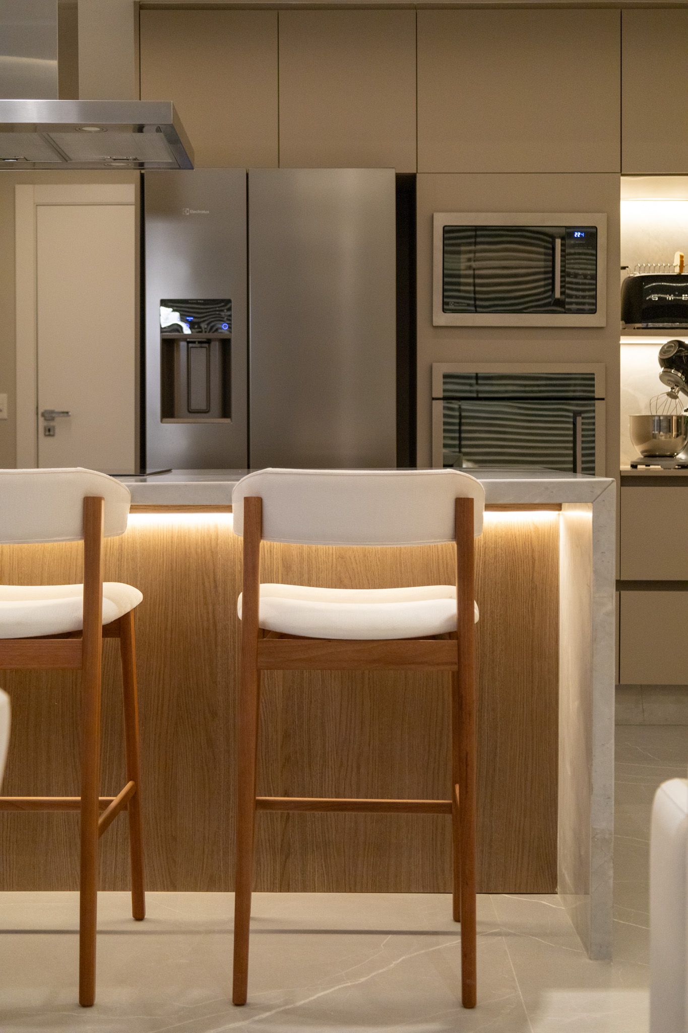 Ilha de cozinha com dois bancos altos; geladeira de aço inoxidável, tons neutros.