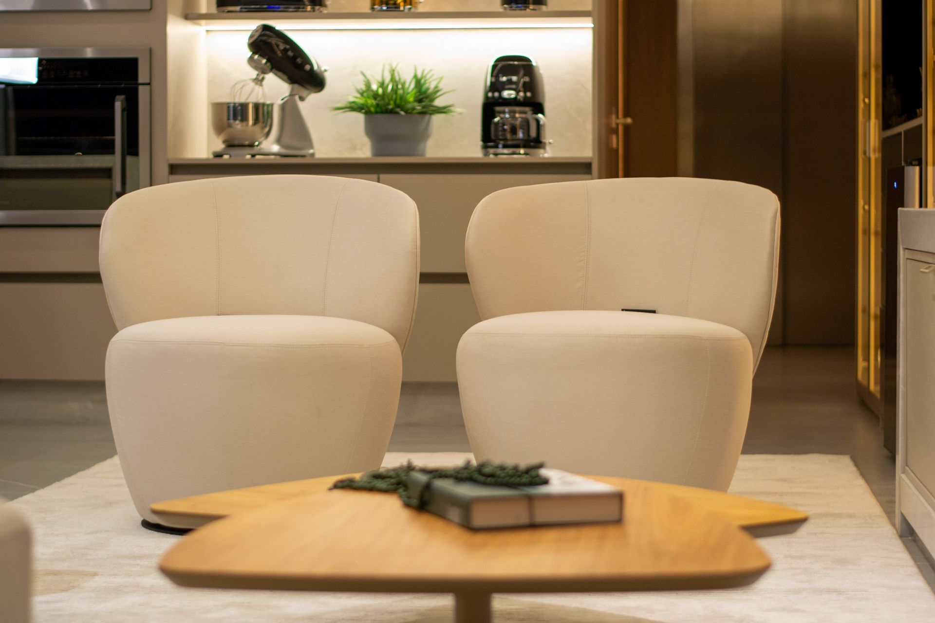 Duas poltronas cor creme estão de frente para uma mesa de centro de madeira com um livro e uma planta. A cozinha aparece ao fundo.