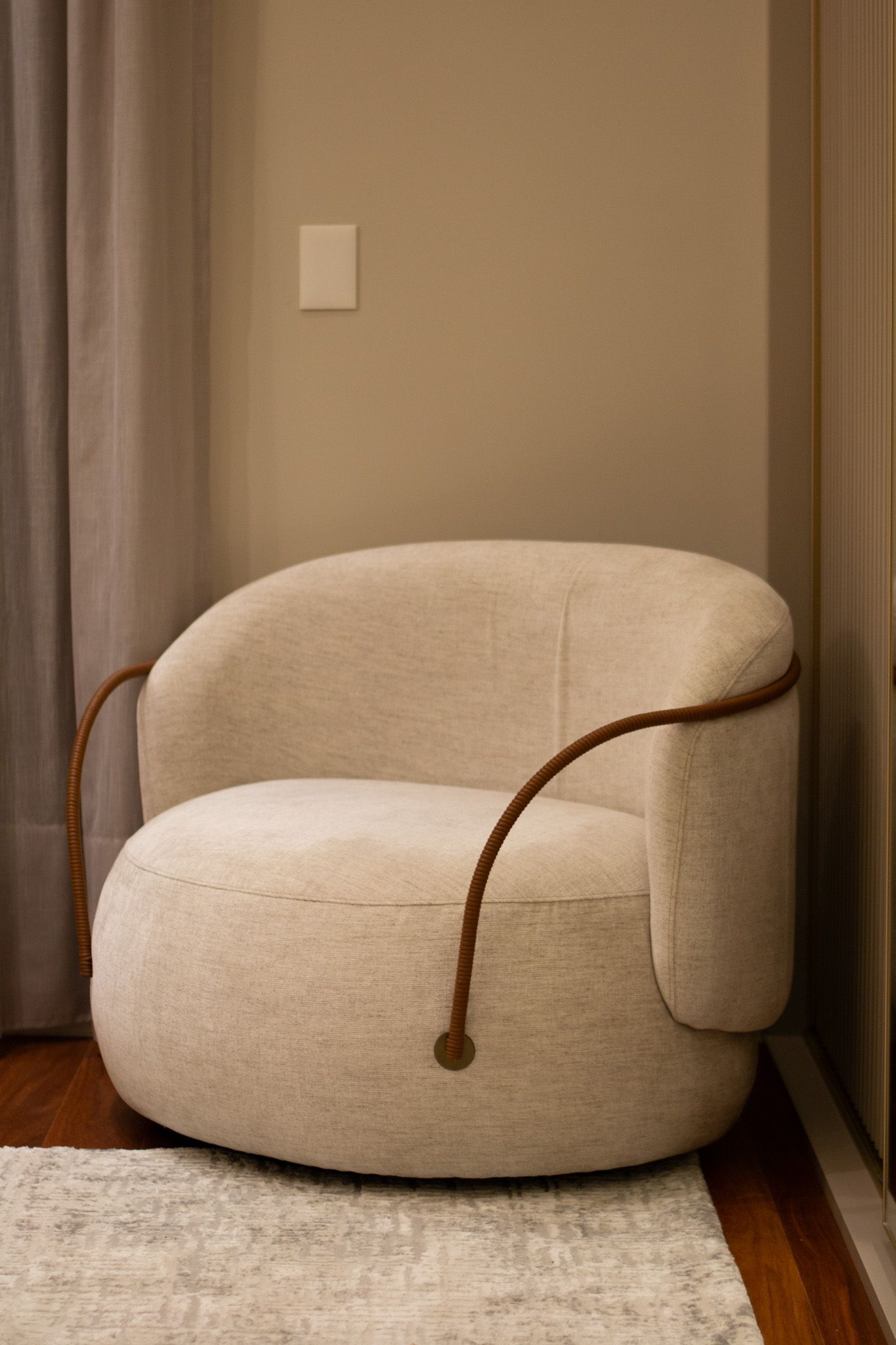 Poltrona bege em tecido bouclé com braços em tom marrom sobre um tapete, encostada em uma parede neutra.