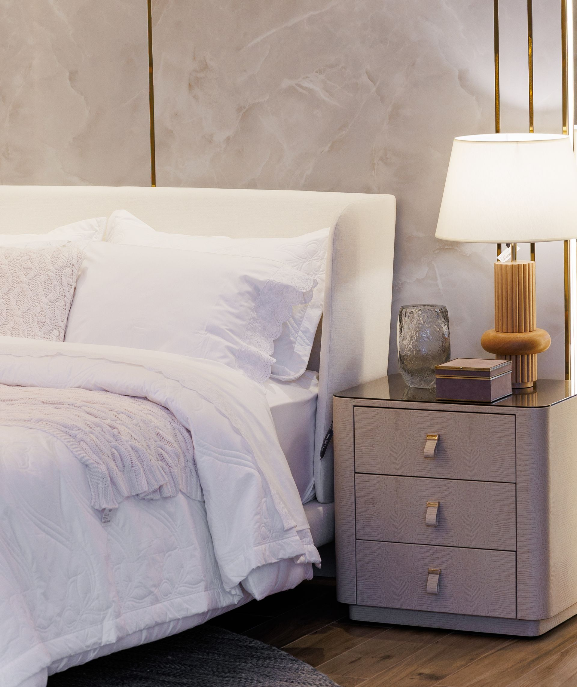 Quarto com cama branca, mesa de cabeceira, abajur e parede texturizada.