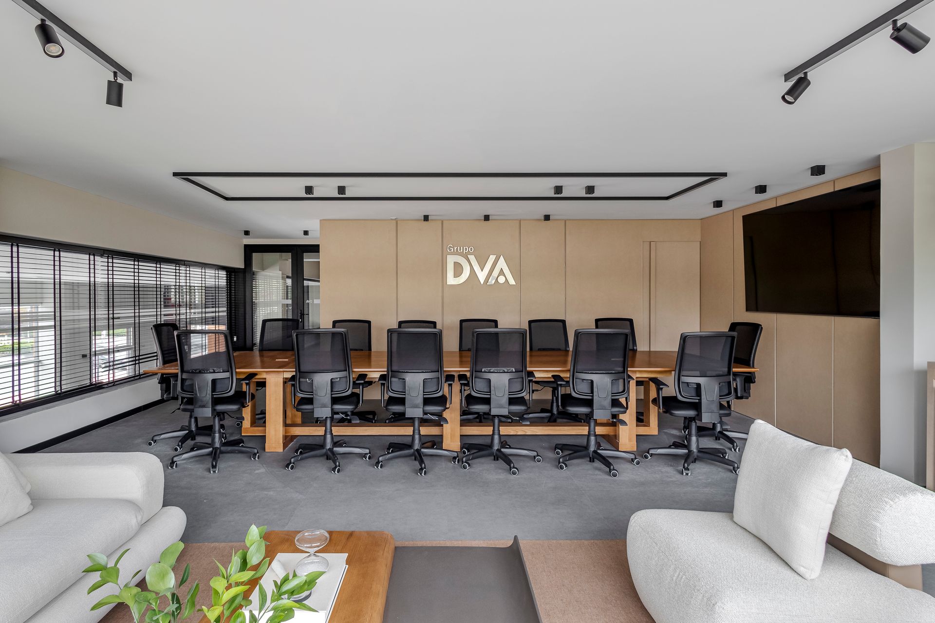 Sala de conferências moderna com mesa longa de madeira, cadeiras pretas, logotipo e dois sofás brancos.