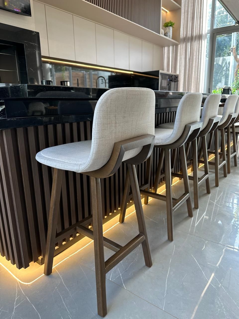 Banquetas de bar enfileiradas em um balcão, com almofadas de cor clara e estrutura de madeira. O balcão tem um tampo escuro.