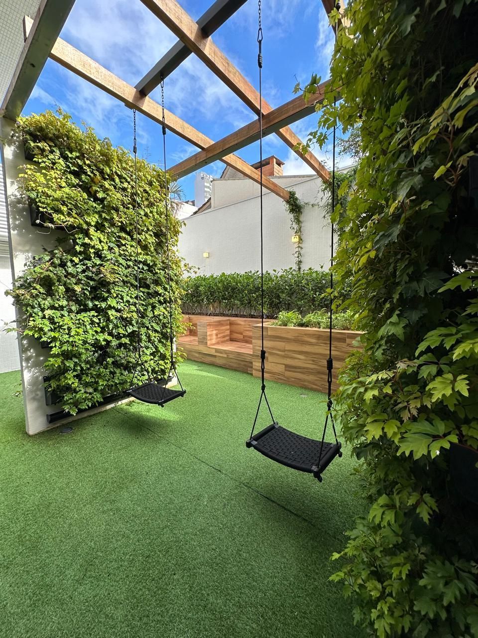 Jardim murado de verde com balanços sob uma pérgola de madeira, grama verdejante e folhagem exuberante.