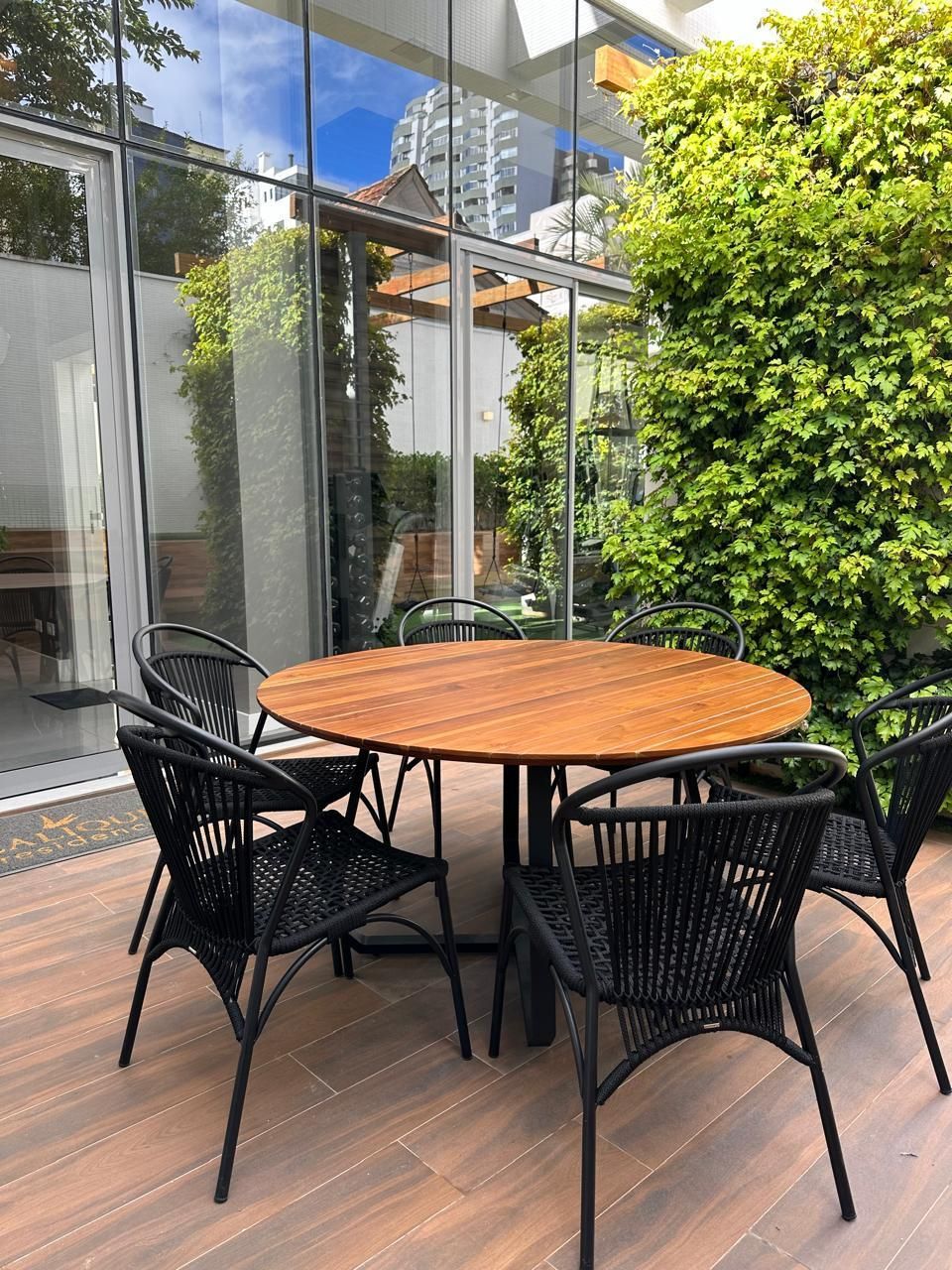 Mesa redonda de madeira e cadeiras pretas em um pátio, com uma parede de trepadeiras verdes.