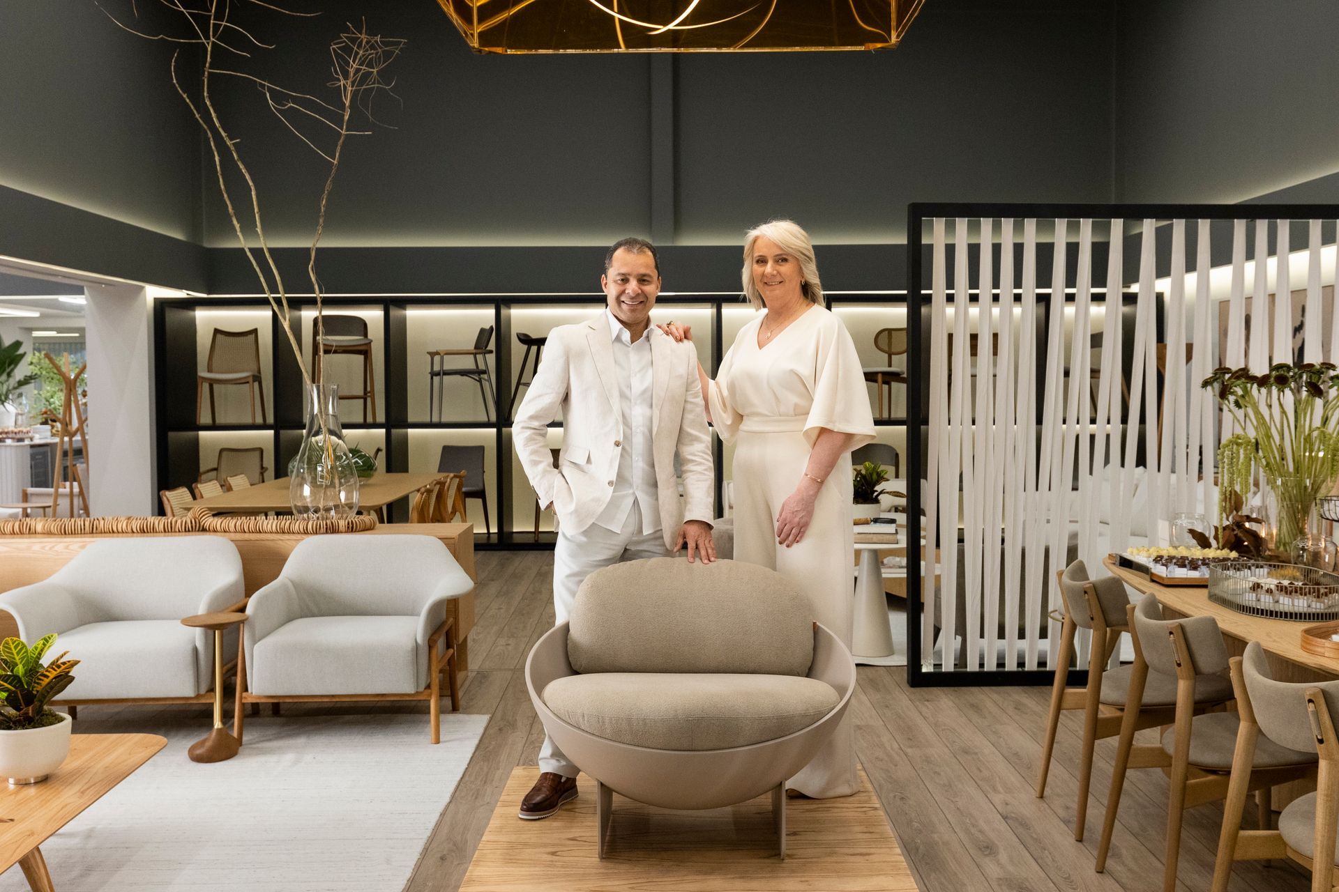 Duas pessoas posam ao lado de uma cadeira bege curva em uma sala de exposição de móveis. Paredes escuras e móveis de madeira.