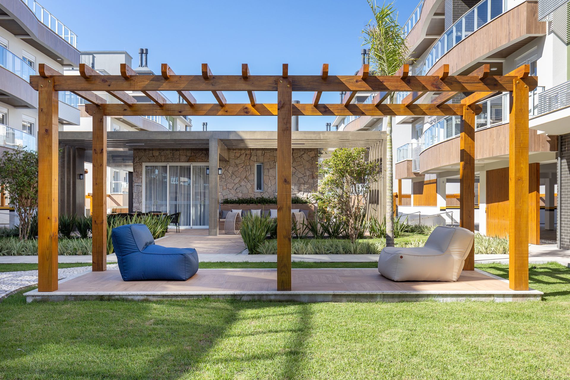 Pérgola de madeira com duas cadeiras tipo pufe em um gramado. Prédios ao fundo.
