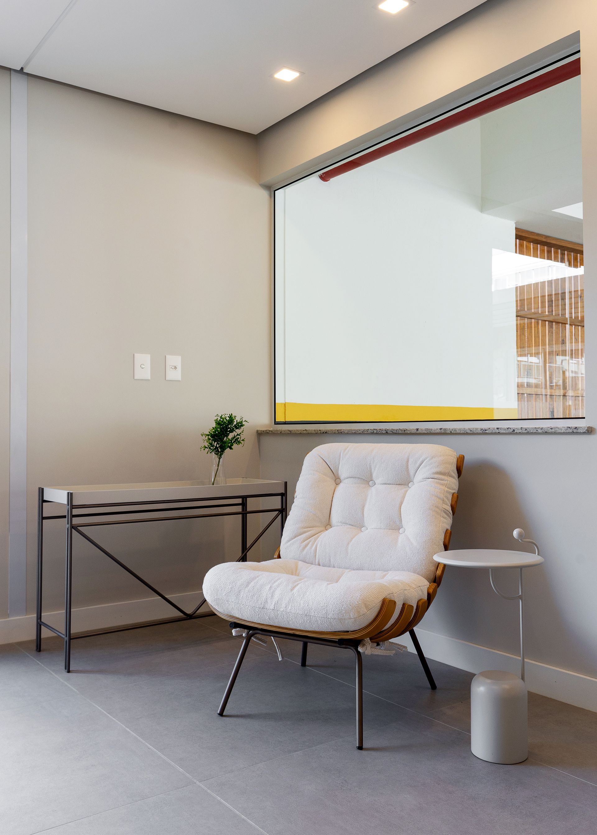 Sala de espera aconchegante com uma cadeira macia, uma mesa pequena e uma janela com vista para outro espaço.