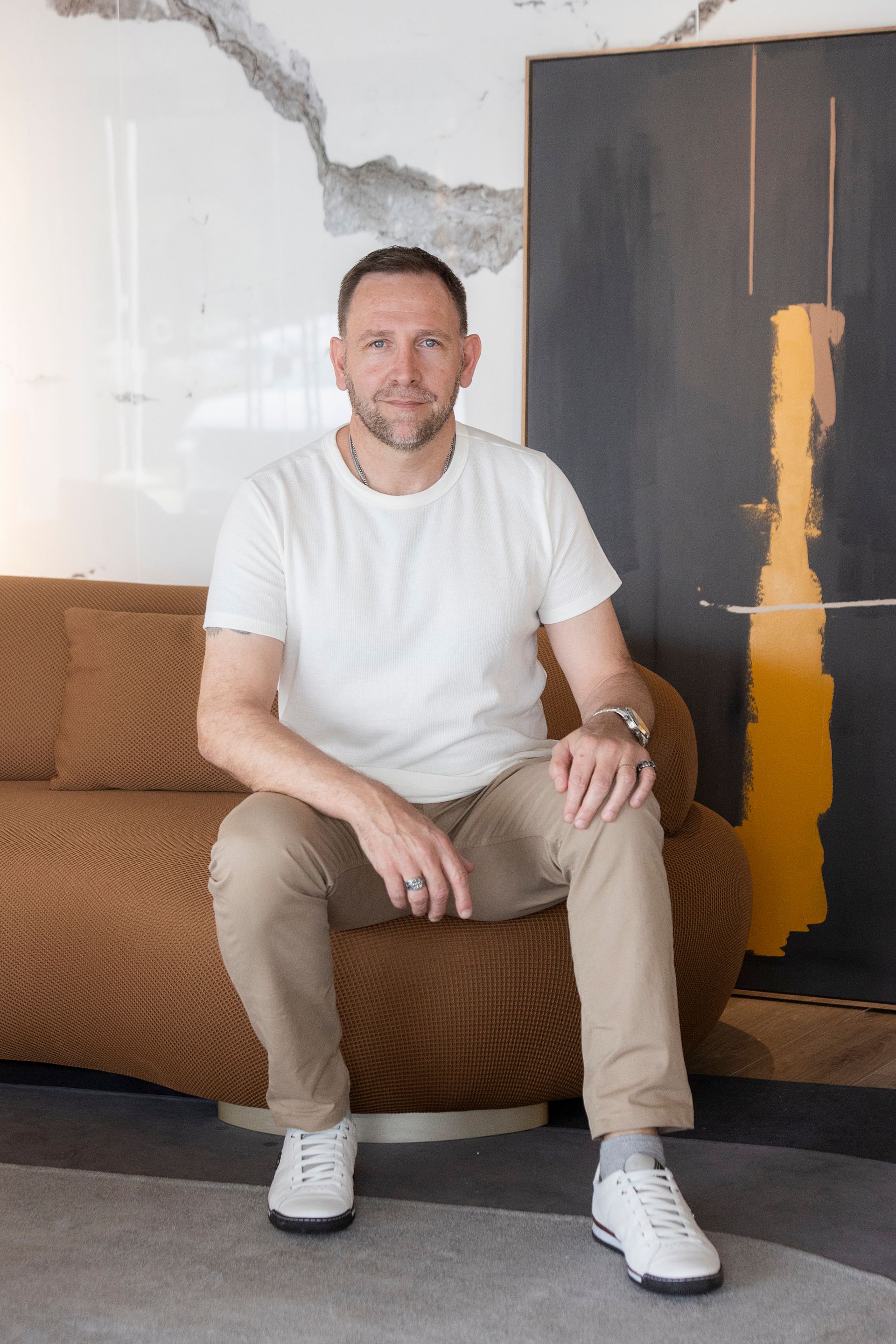 Homem sentado em um sofá marrom, vestindo camisa branca, calça bege e tênis brancos. Obra de arte na parede.