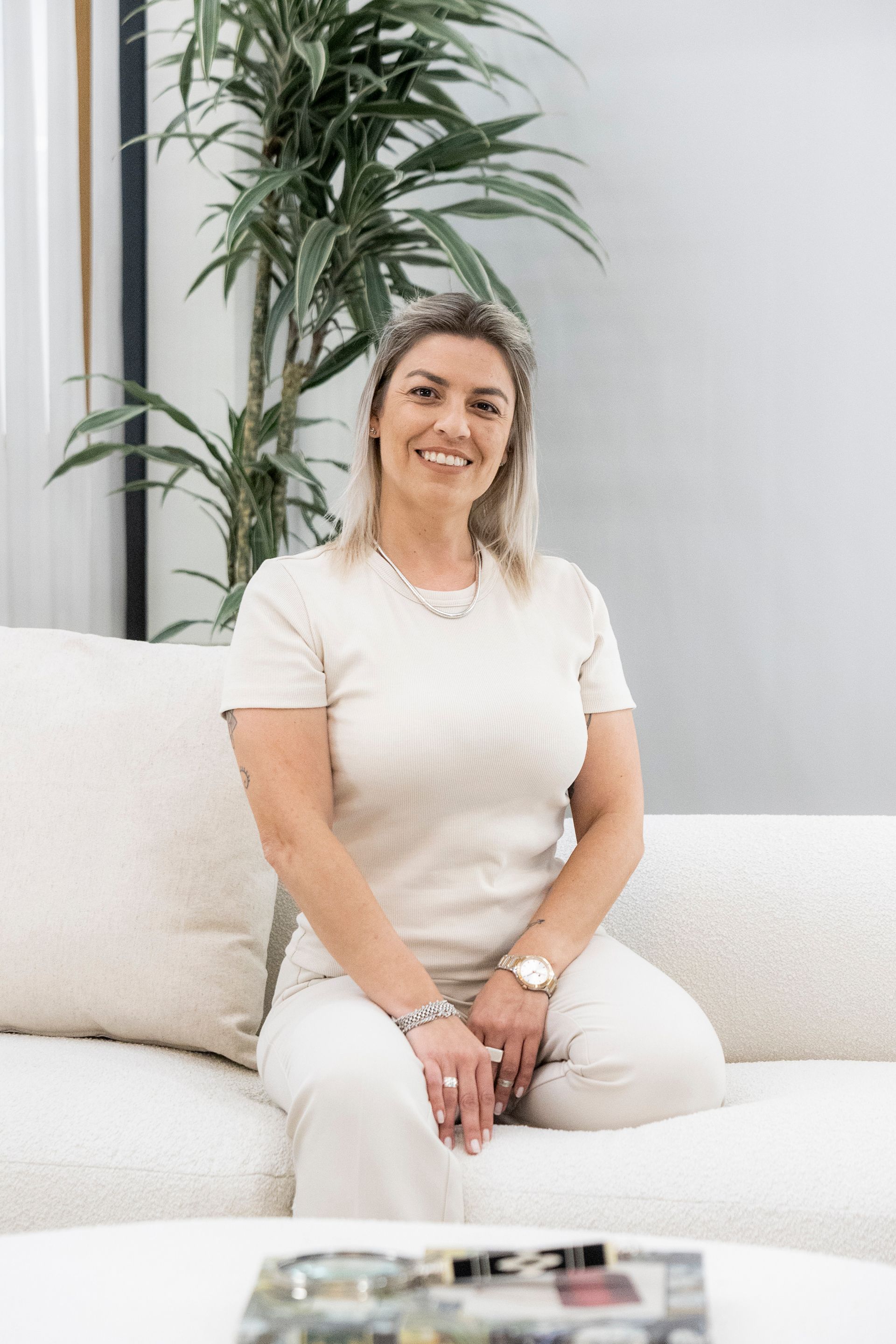 Mulher loira sentada em um sofá branco, sorrindo, vestindo uma roupa creme. Planta verde ao fundo.