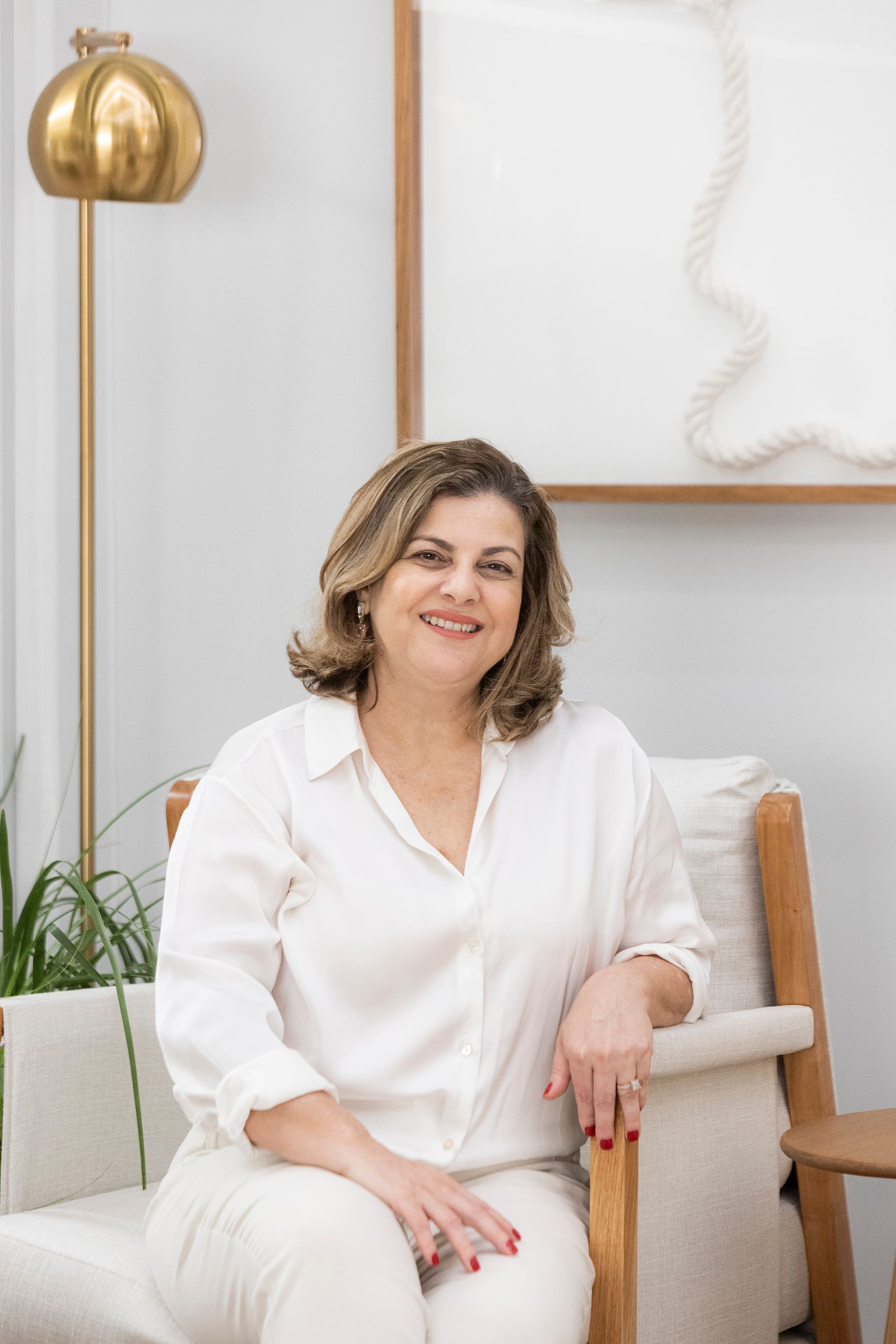 Mulher de camisa branca sentada em uma cadeira clara, sorrindo para a câmera em uma sala iluminada.