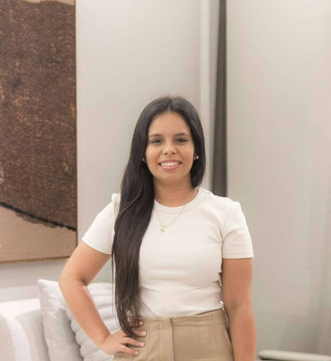 Mulher com longos cabelos escuros, sorrindo, vestindo uma camisa branca e calças bege, em uma sala bem iluminada.