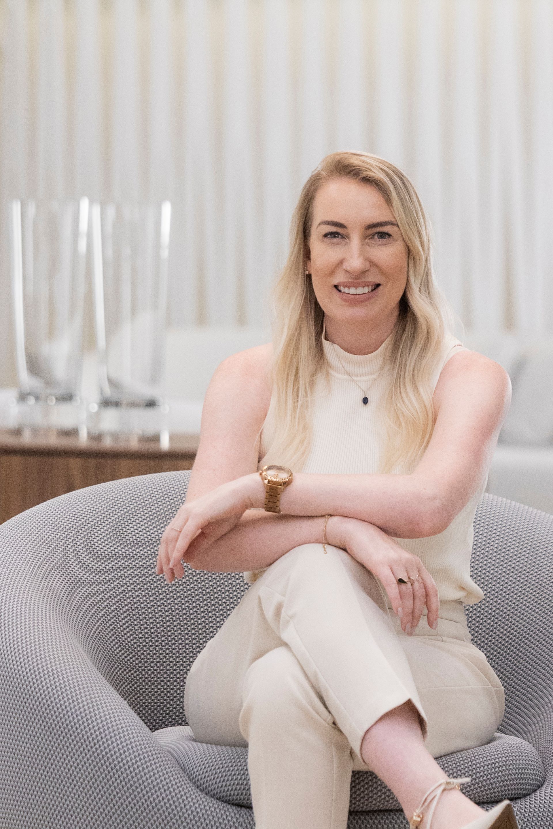 Mulher loira com roupa creme sentada em uma poltrona cinza, sorrindo, braços cruzados, em uma sala de estar.