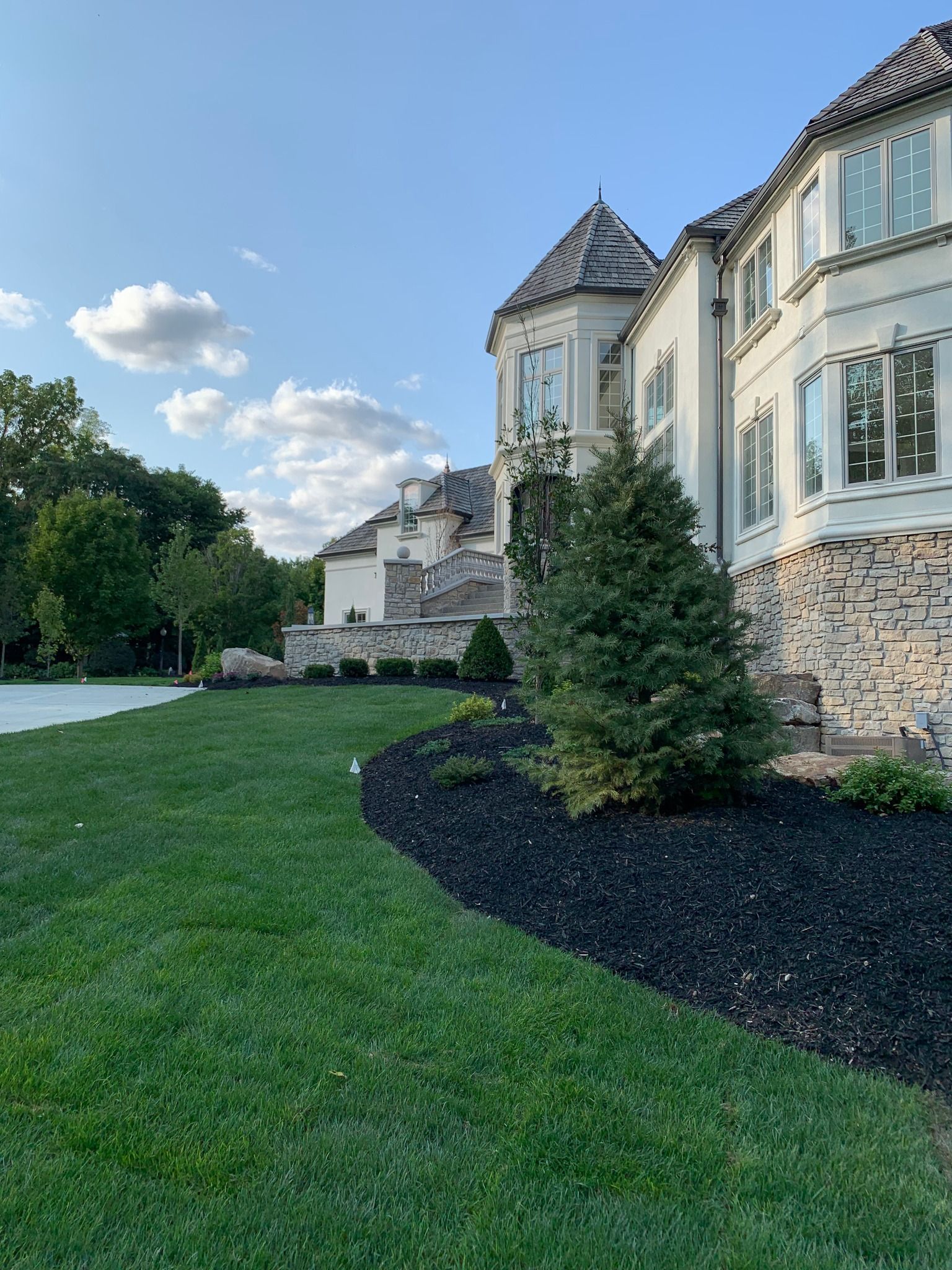 A large, light-colored house with a tower and stone facade, black mulch and green grass lawn.
