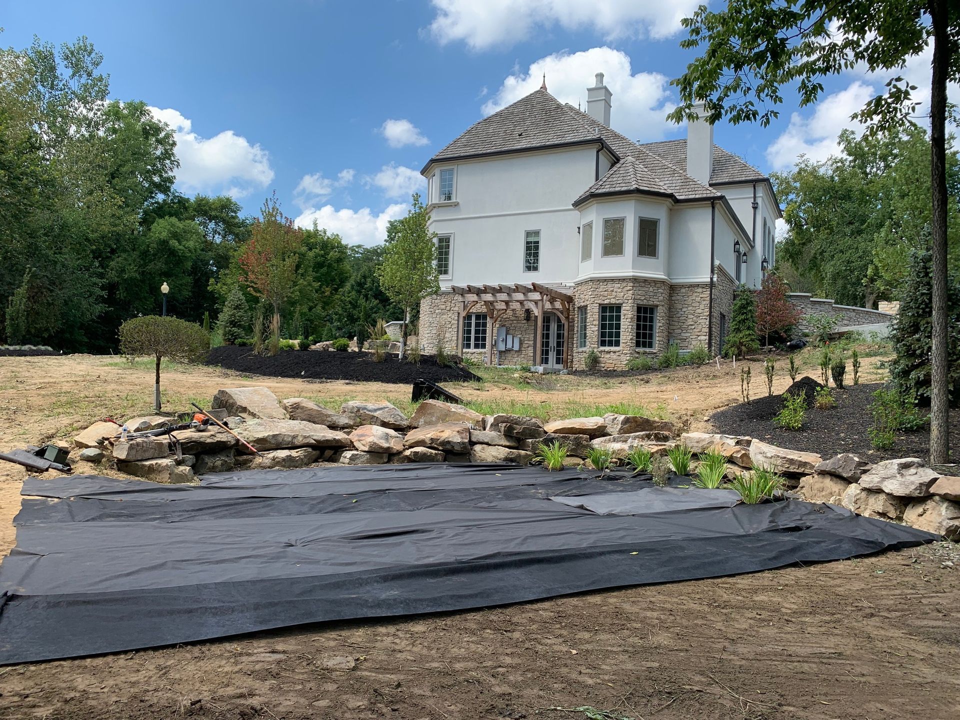 Large white house with stone accents, a dark tarp in the foreground, and landscaping in progress under a blue sky.