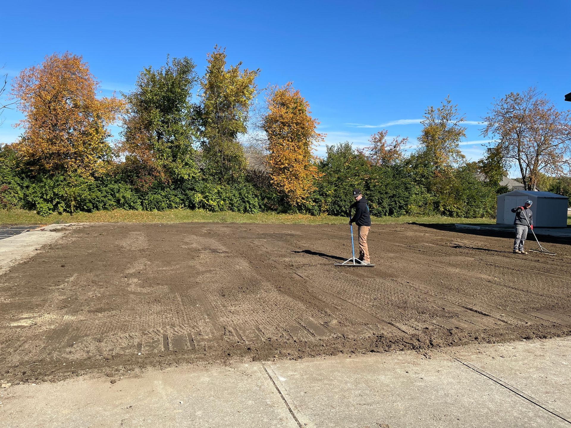 People raking soil in a cleared area, preparing for planting, trees with fall foliage in the background.