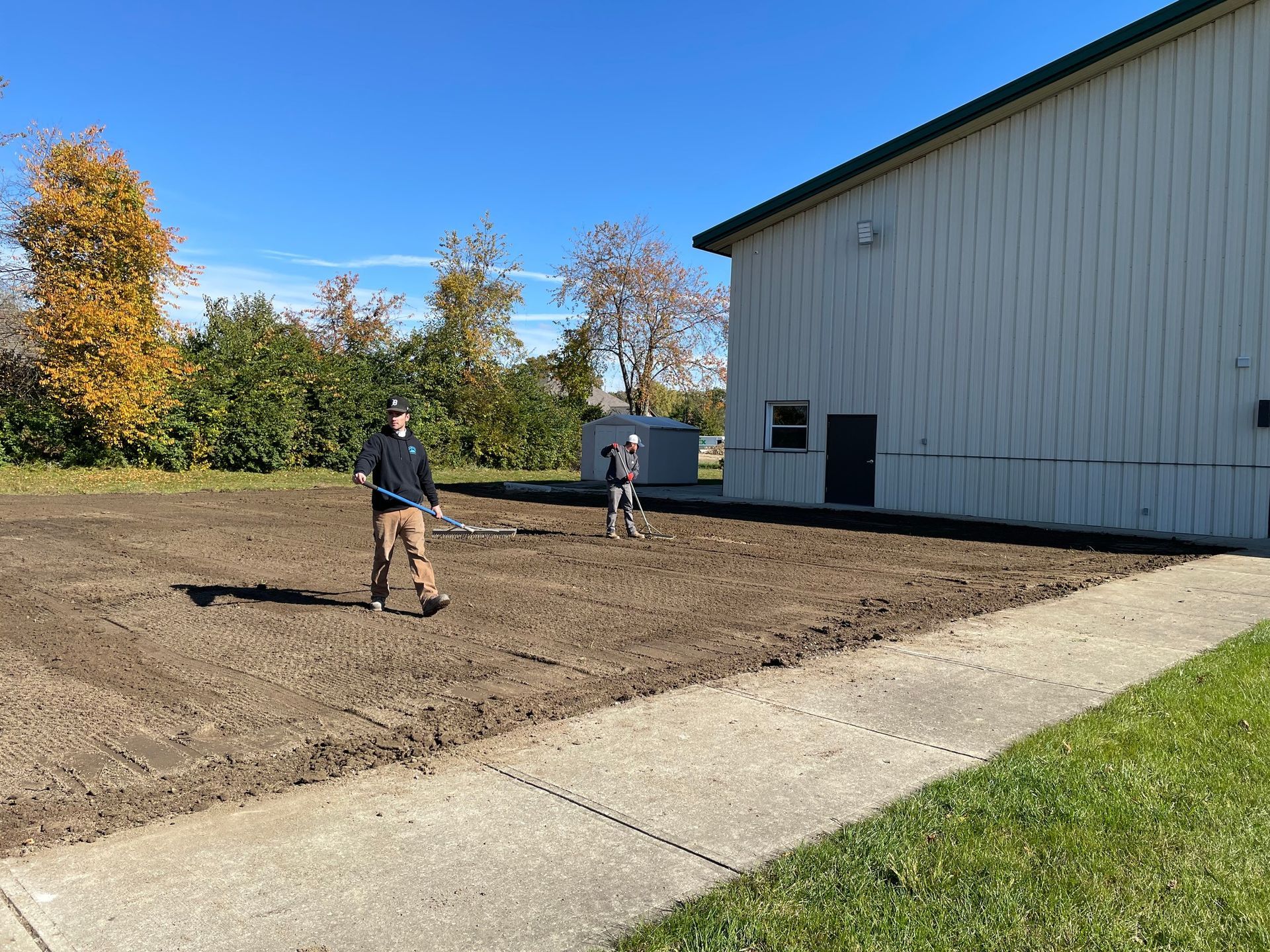 Two people raking soil next to a building on a sunny day.