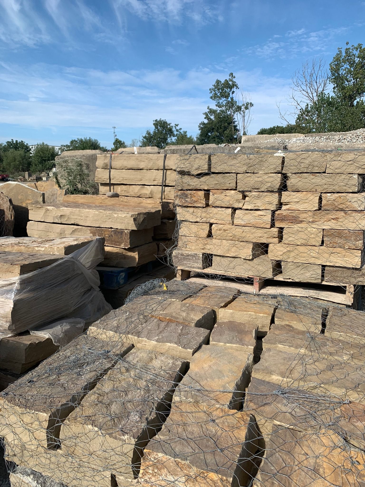 Stacks of rectangular stones, various shades of tan and brown, on pallets, outdoors. Blue sky with clouds.