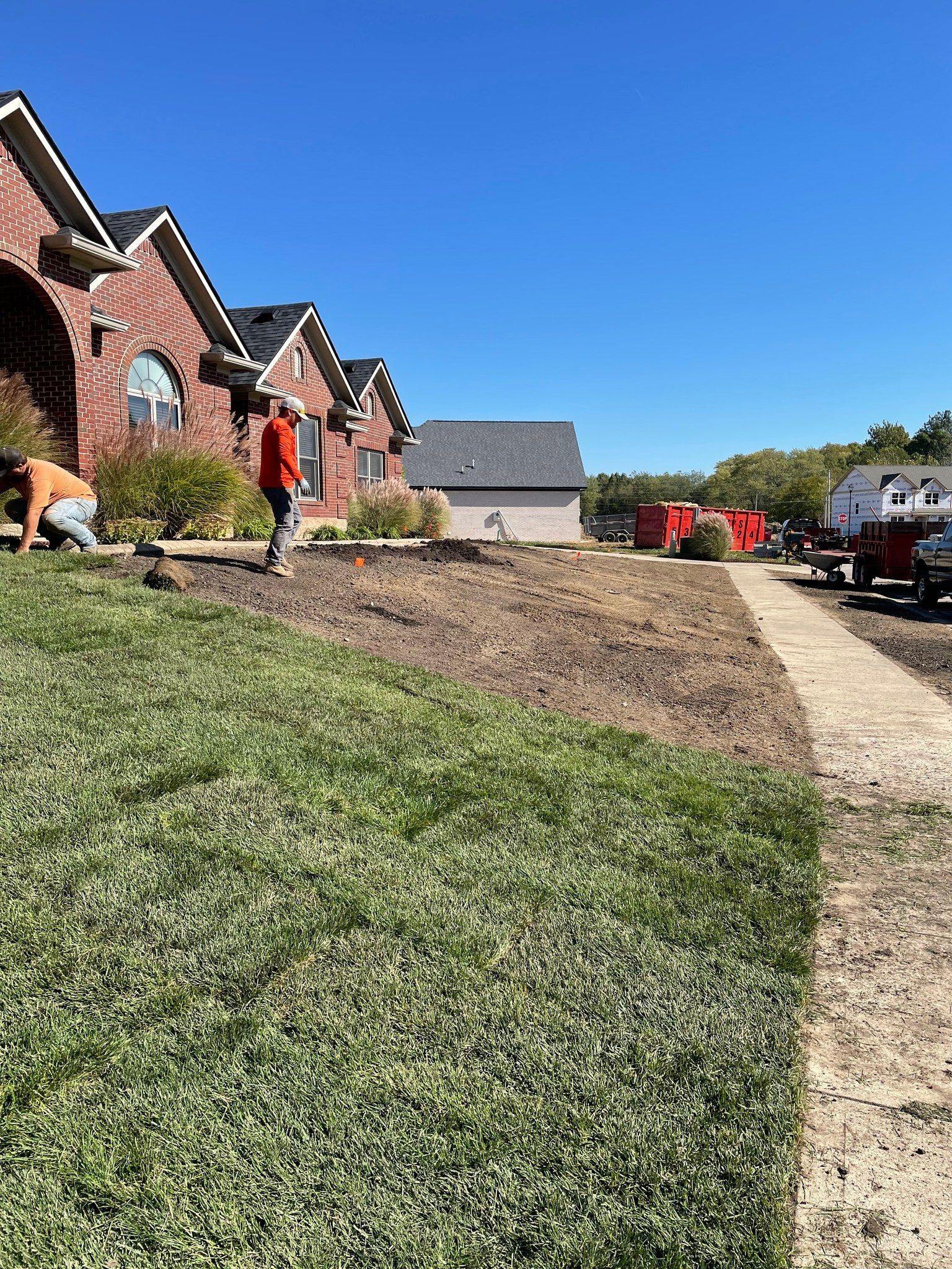 Workers landscaping a lawn in front of a brick house under a clear blue sky.