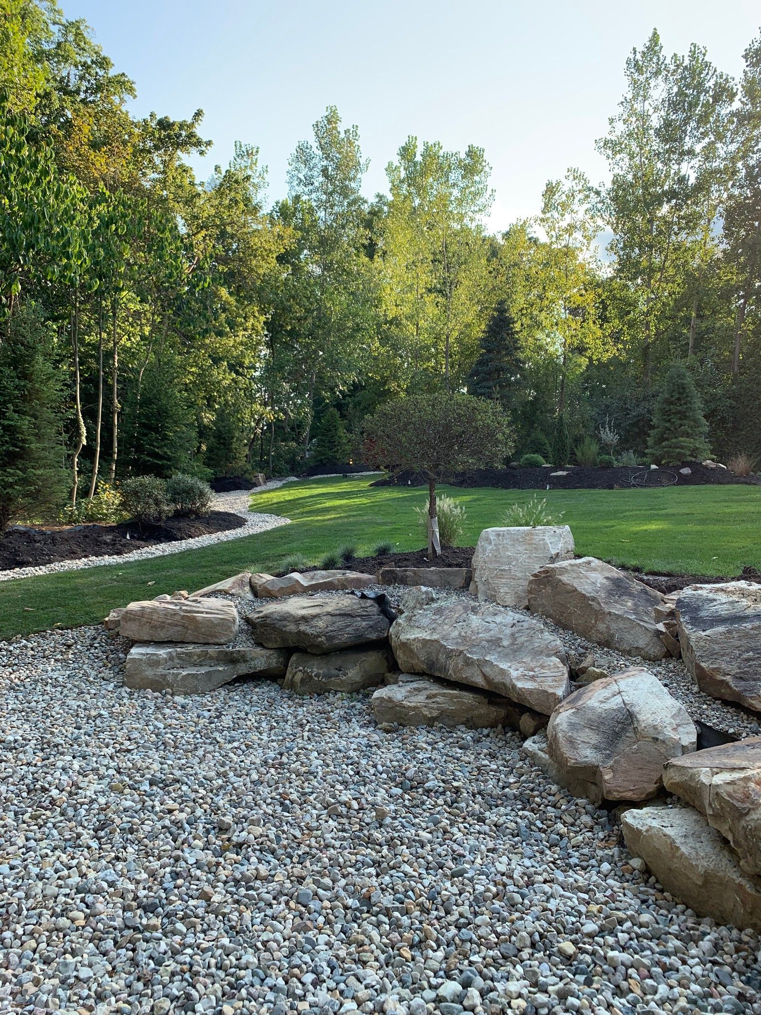 Rock wall and gravel in front of a grassy lawn and trees under a sunny sky.
