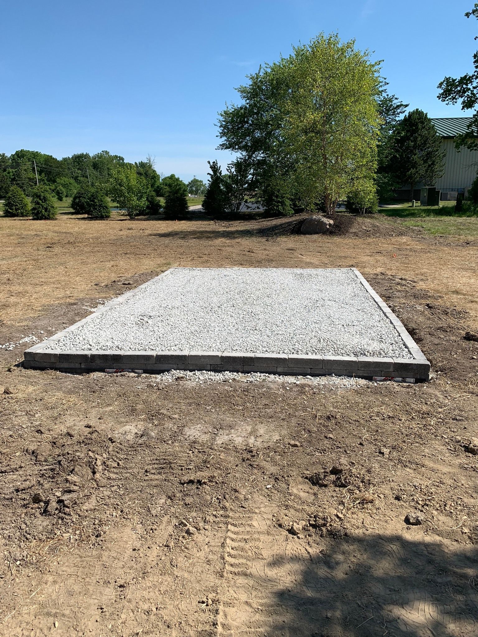 Gravel-filled rectangular foundation on dirt, with trees and a building in the background under a blue sky.