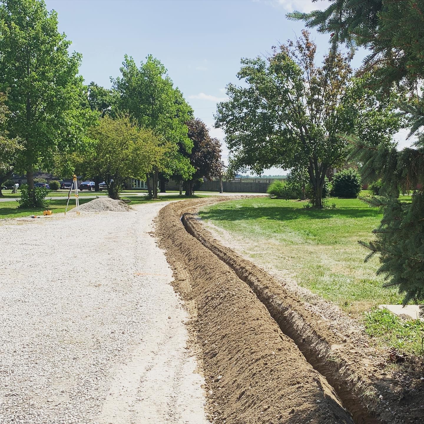 A trench dug in gravel and grass, trees in the background, sunny day.