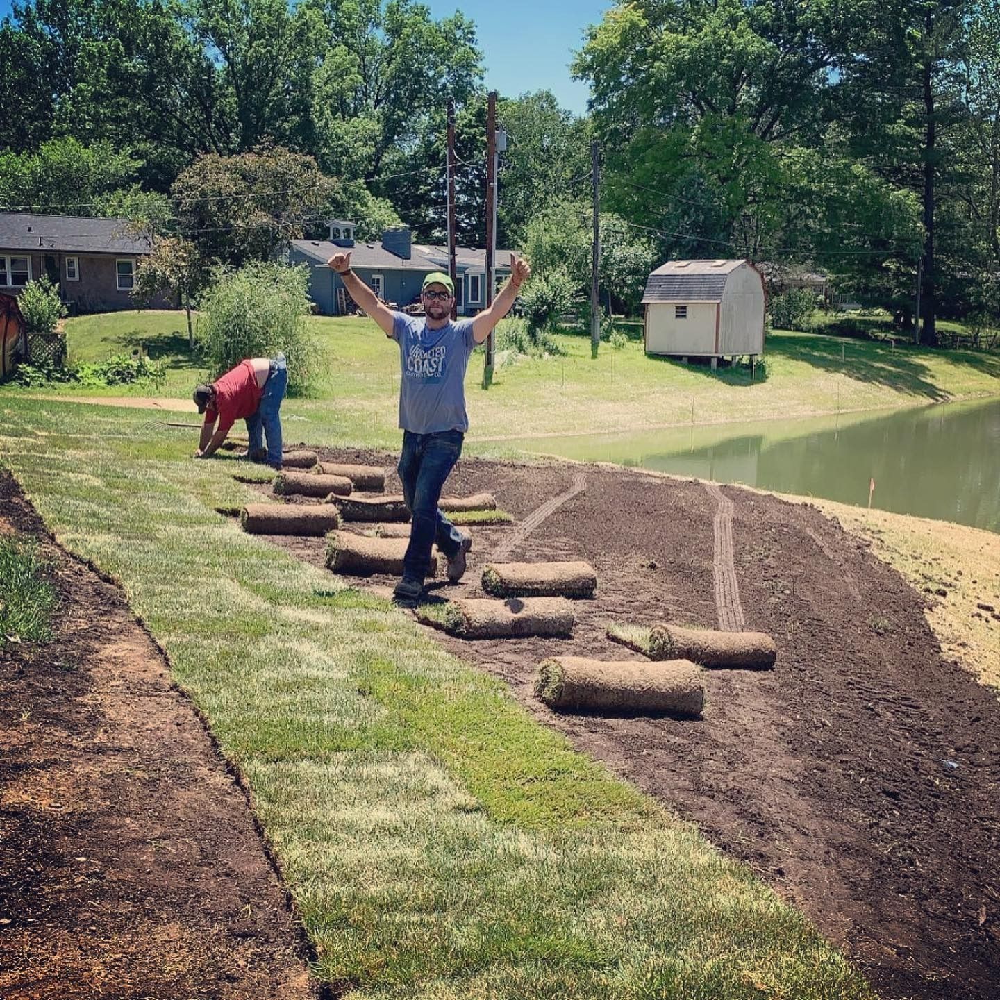 Man celebrates laying new sod rolls near a pond. Others work in the background on a sunny day.