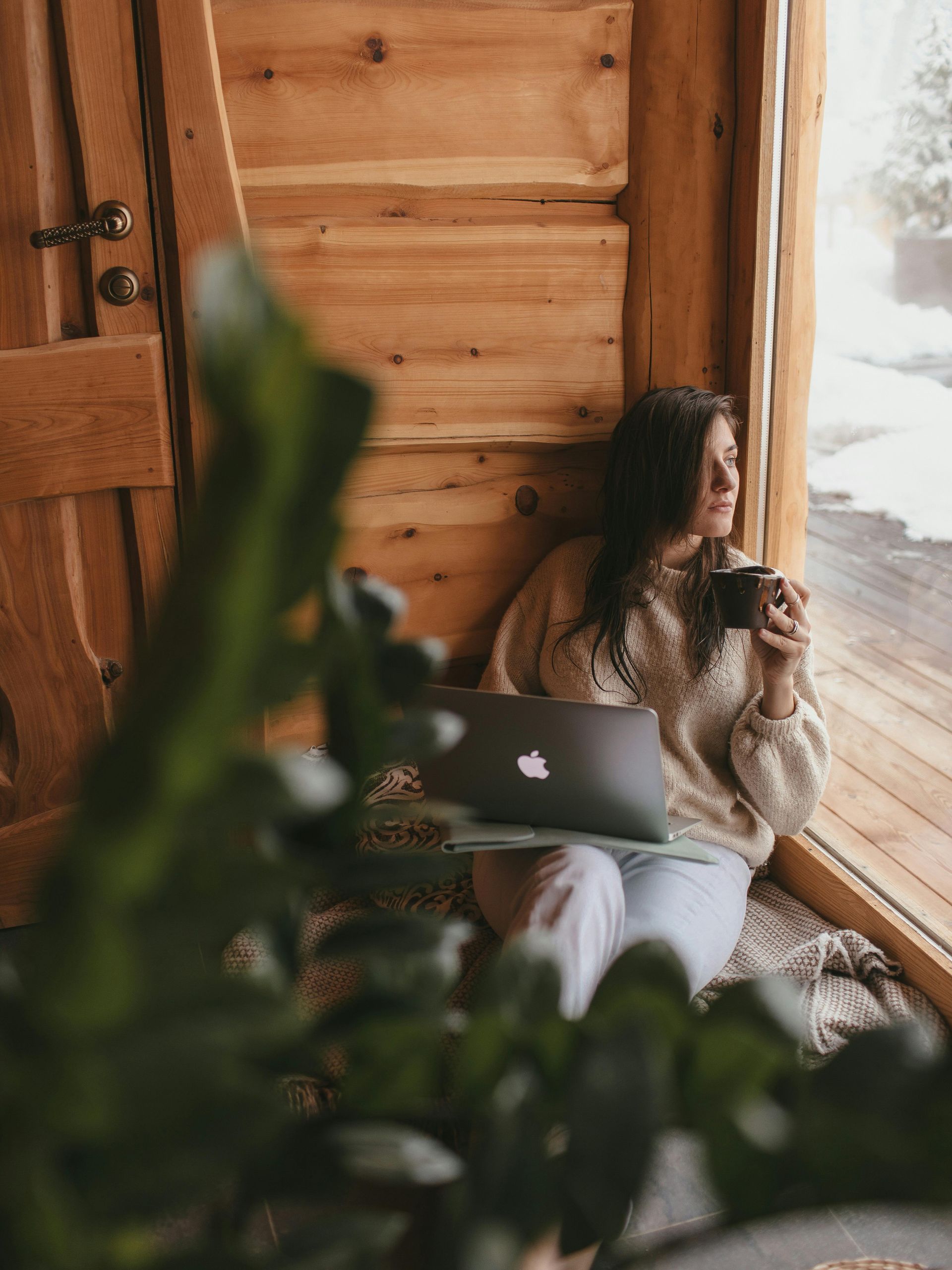 Una mujer está sentada en el alféizar de una ventana con una computadora portátil y una taza de café.