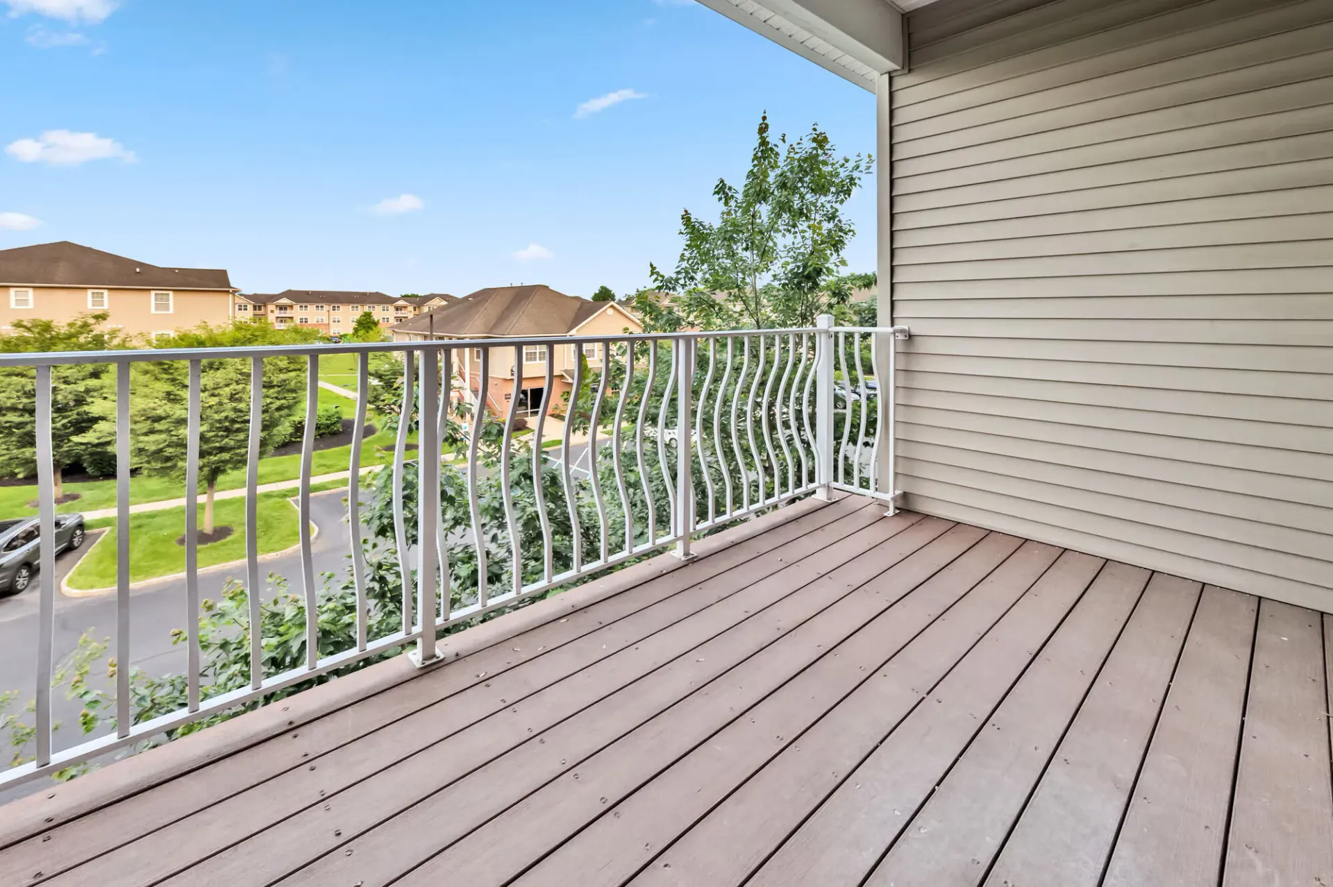 Balcony of an apartment with a wooden deck and white railing, overlooking trees and nearby buildings.