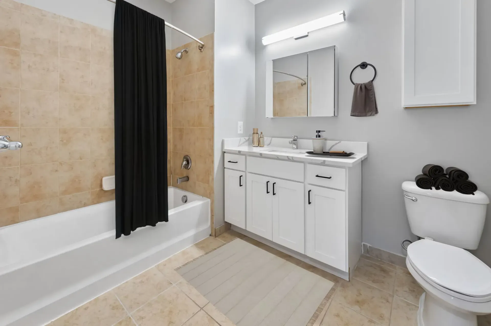 Modern apartment bathroom with beige tile tub/shower surround, black shower curtain, white vanity, and toilet.