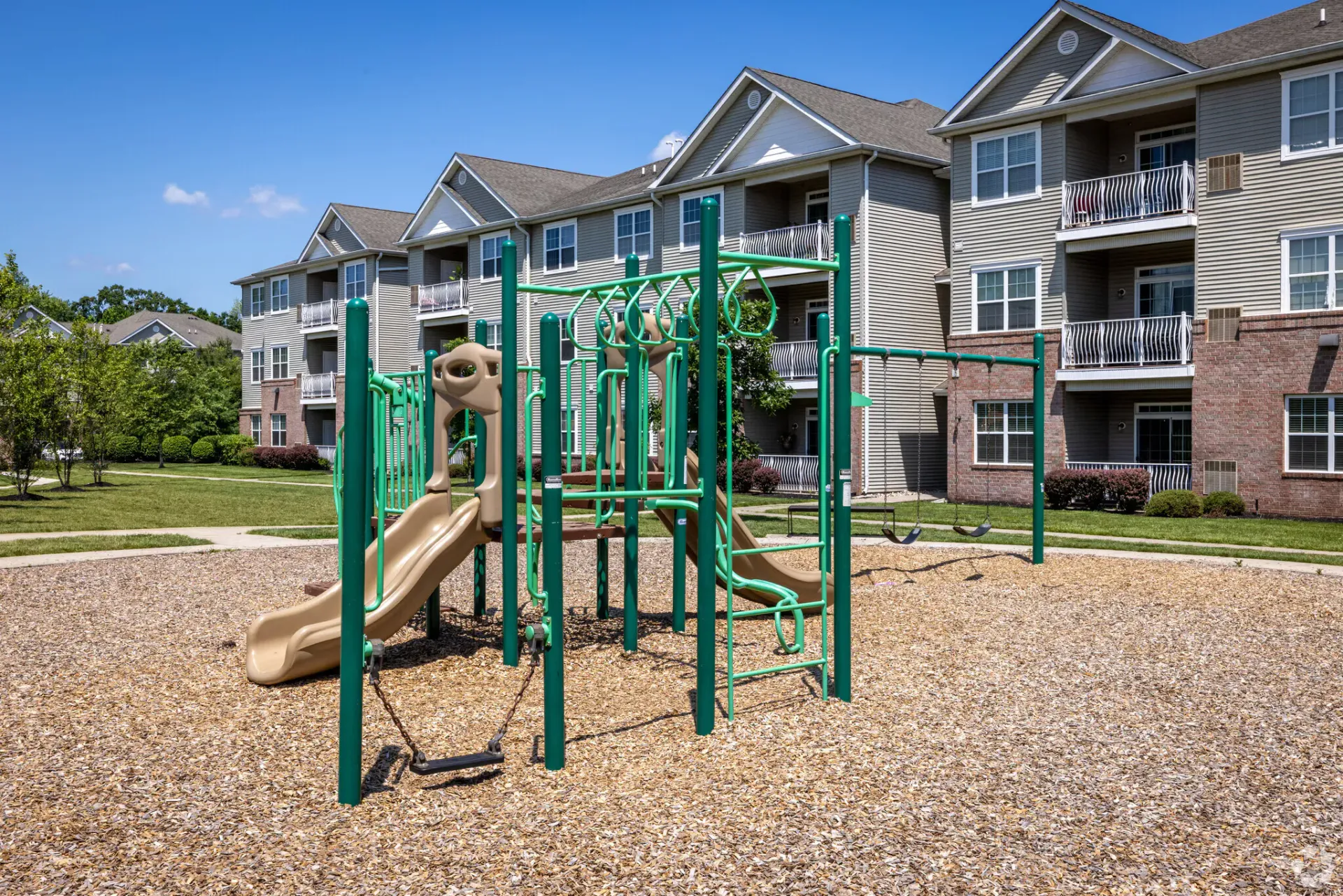 Green playground with a slide, climbing structures and swings in a courtyard of apartment buildings.