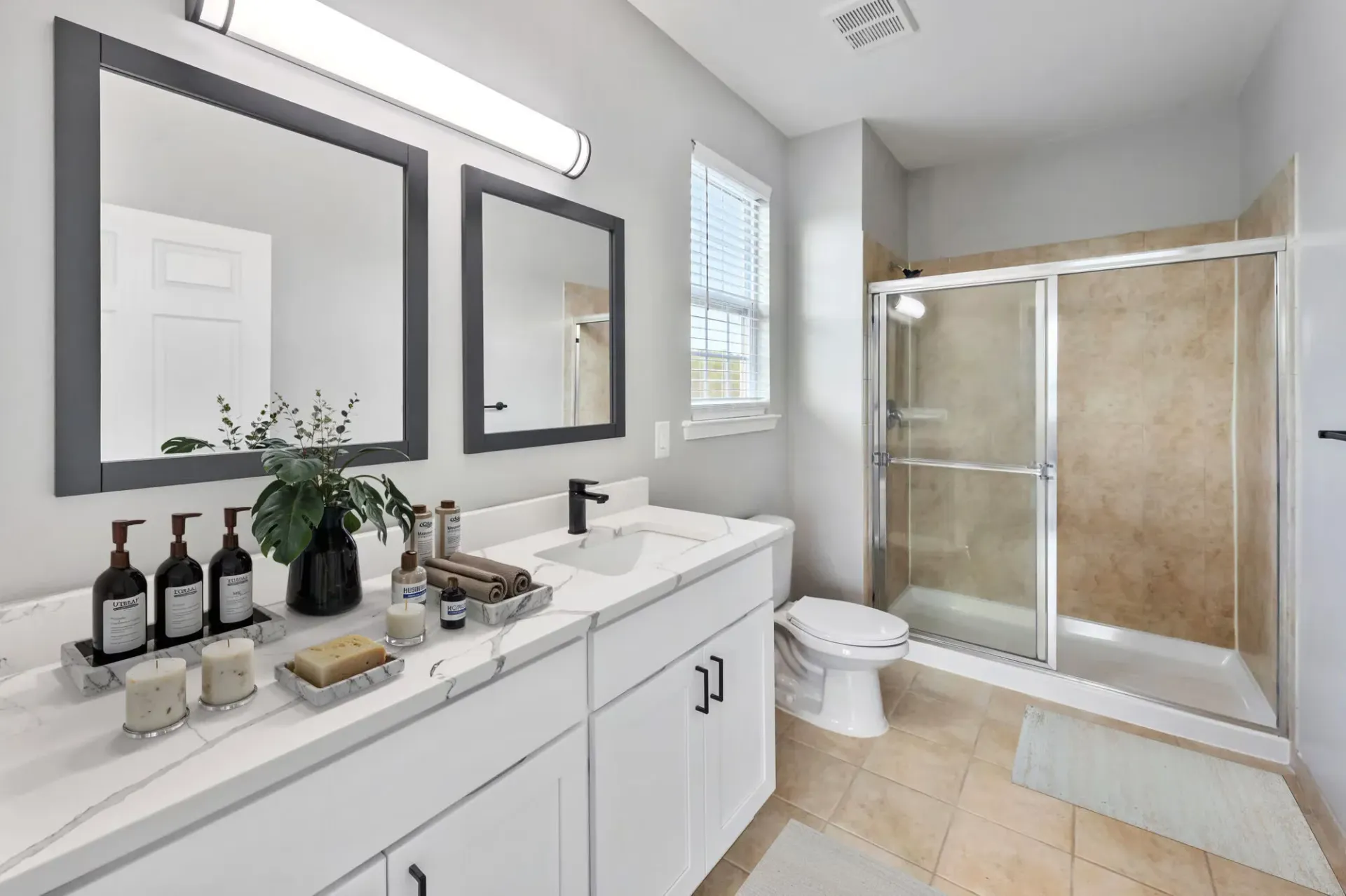 Bathroom with a white double vanity, two framed mirrors, a window, and a glass-enclosed shower.