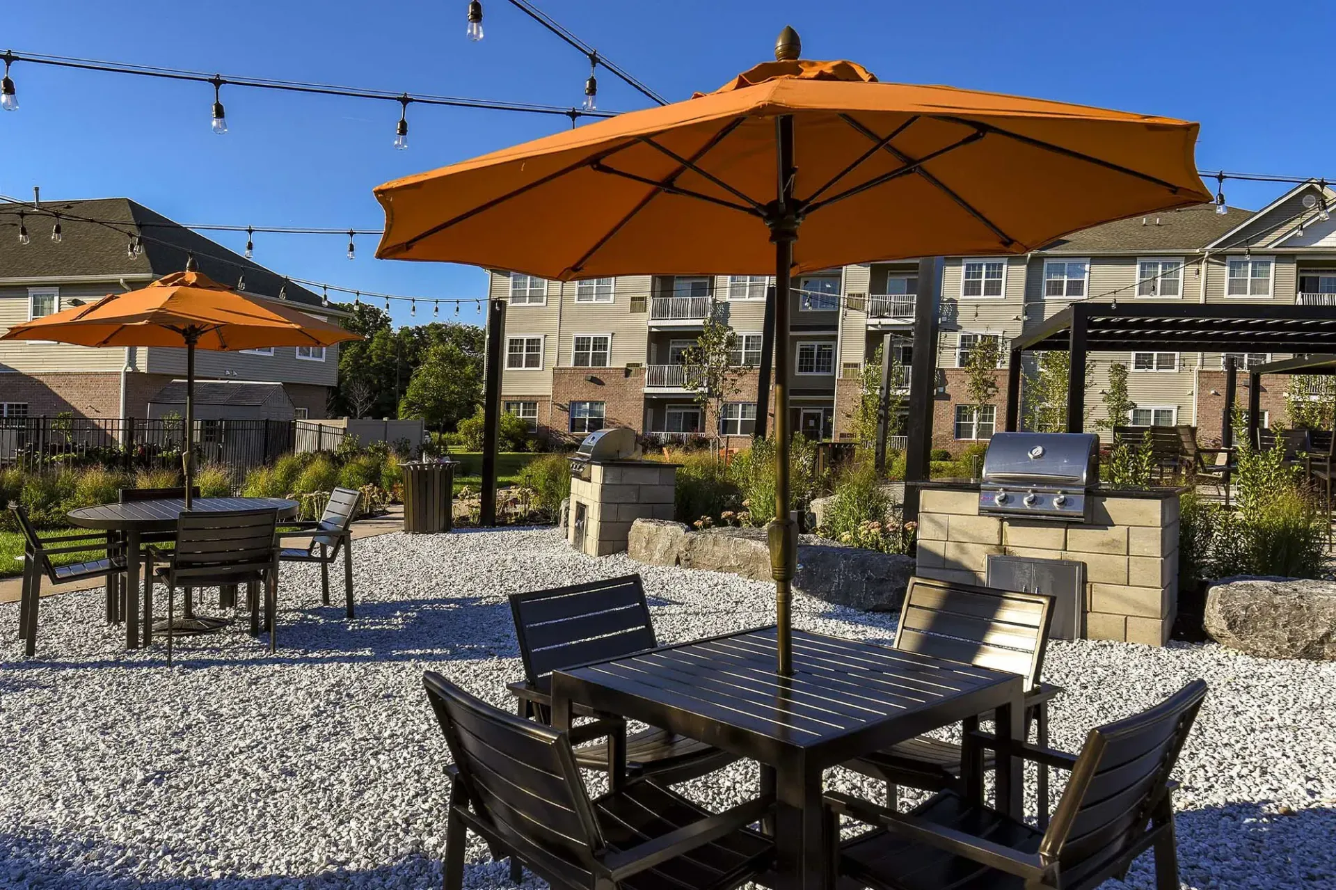 Outdoor community courtyard with orange umbrellas, string lights, gravel ground, and seating.