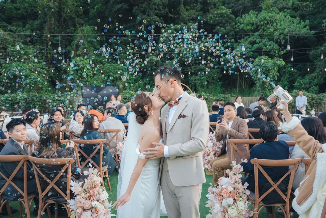 Newlyweds kiss at outdoor wedding ceremony; bubbles float above guests seated in rows.