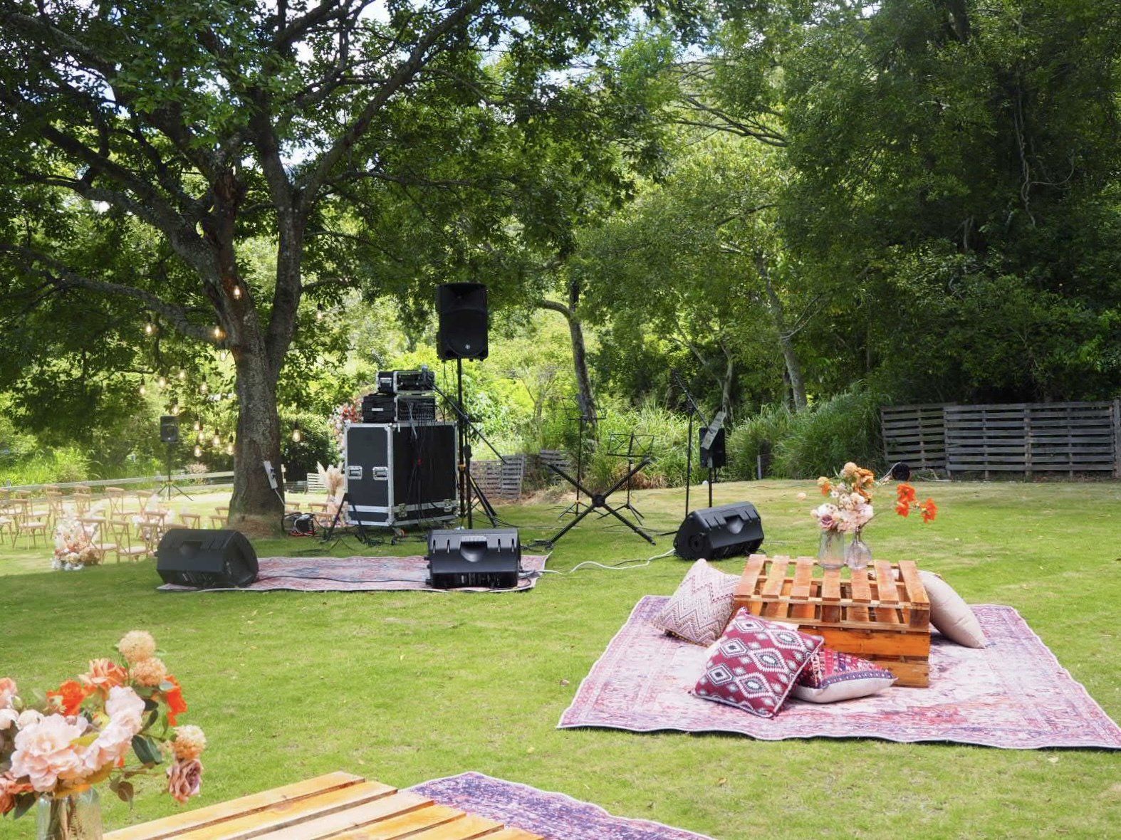 Outdoor event setup with sound equipment, rugs, and seating in a grassy area, under trees.