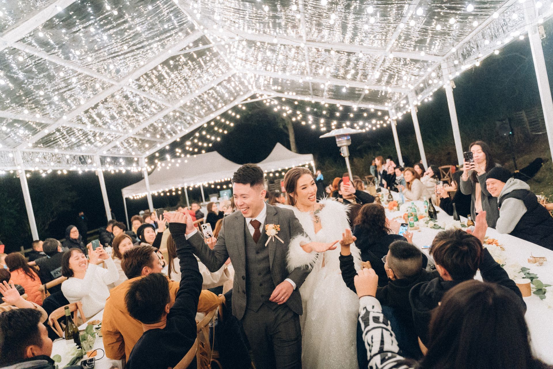 Newlyweds walk through guests, under a canopy of lights at a wedding reception.