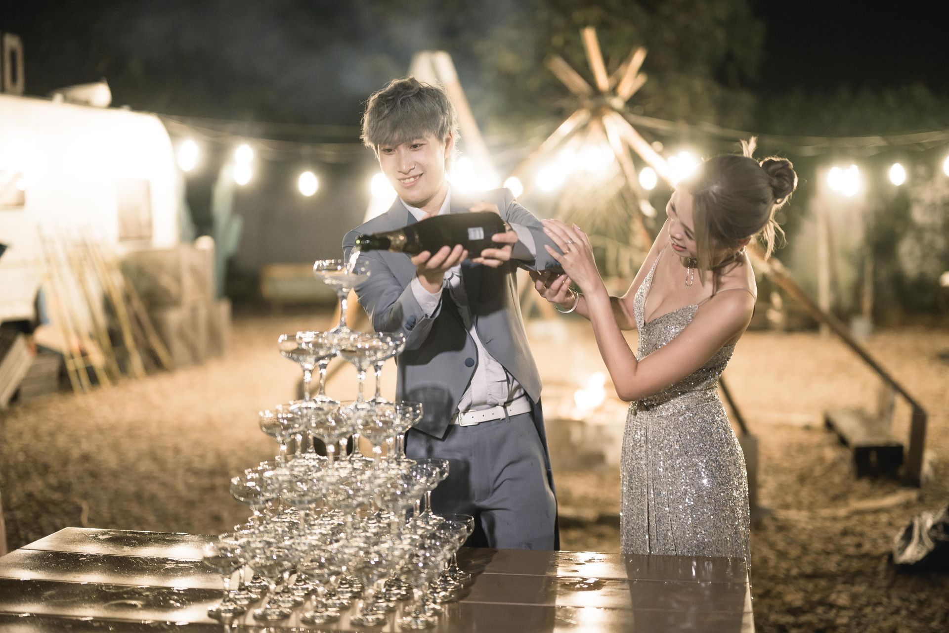 Man pouring champagne into a tower of glasses for a woman in an outdoor nighttime setting, fairy lights visible.