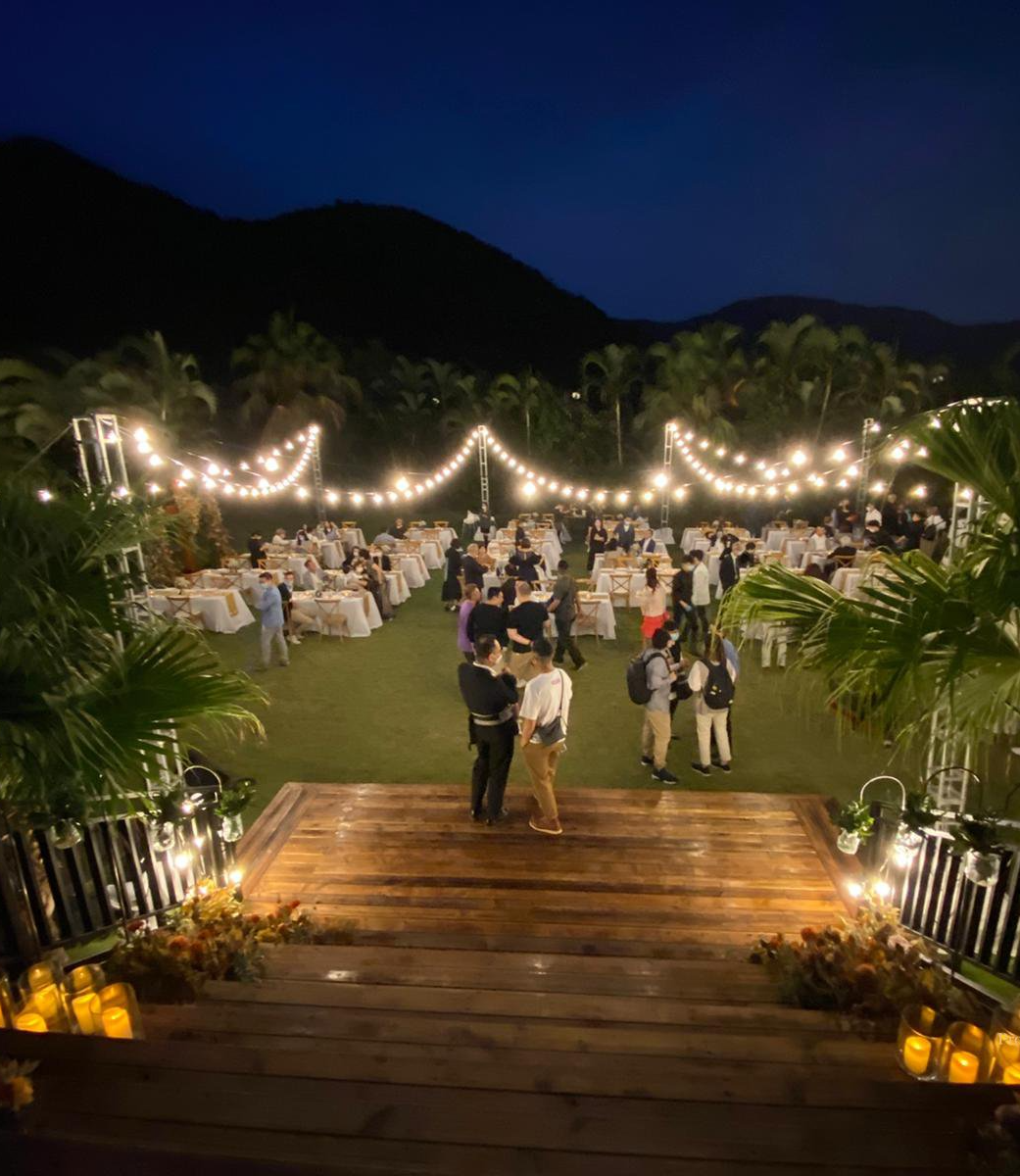 Nighttime outdoor event with tables, string lights, and a wooden walkway. People gather in a lush green area, mountains in the background.