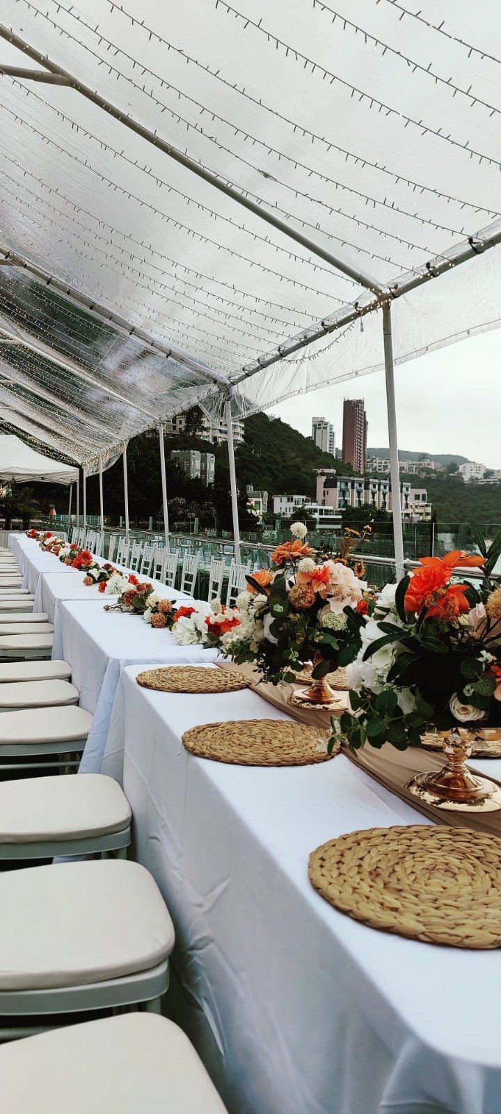 A long table set for a special occasion, under a clear tent, with floral arrangements and city views in the background.