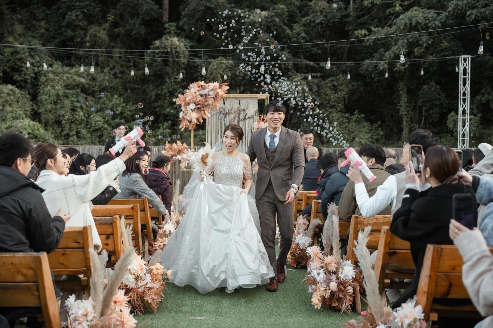 Newlyweds walk down the aisle, cheered on by guests. Outdoor wedding with floral decor.