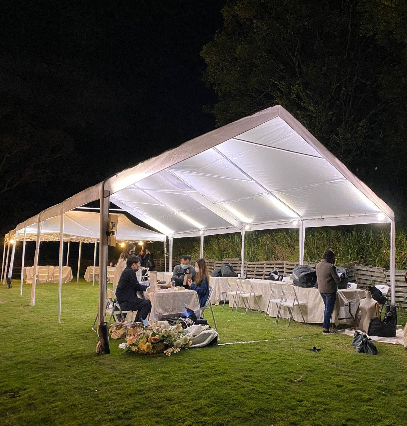 Outdoor event tent at night, people seated around tables, lit up with overhead lights on a grassy lawn.