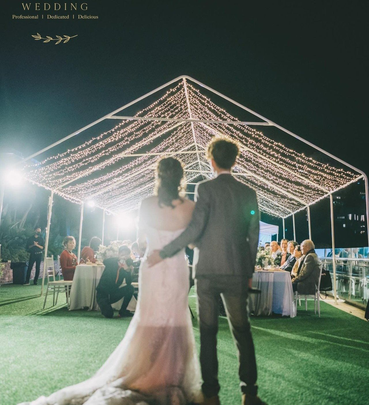 Couple at wedding, arm in arm, gazing at guests under lit canopy at night.