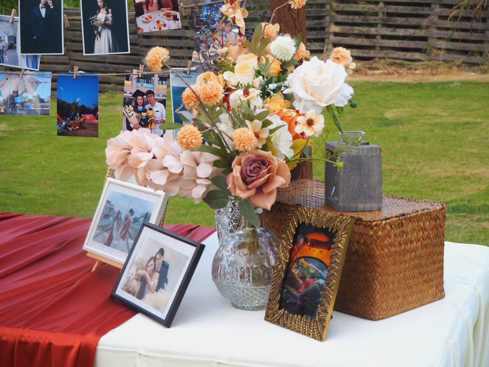Wedding decorations with flowers, photos, and a red cloth on a white table in an outdoor setting.