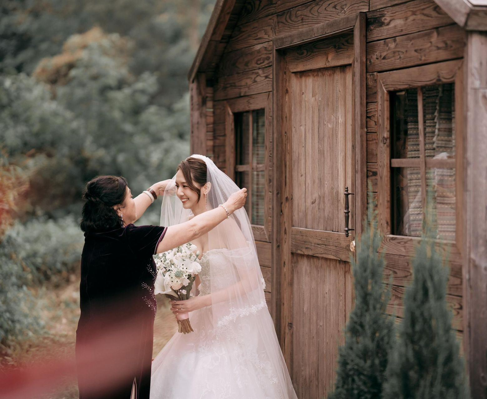Woman in wedding dress and veil, holding bouquet, with another woman adjusting the veil near wooden shed.